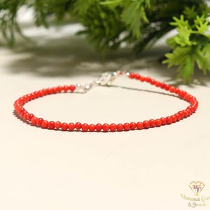 May include: A fine red coral bead bracelet with a silver clasp. The bracelet features small, round, bright red beads. The clasp is a simple, secure design. The bracelet is shown on a light surface with green foliage in the background. A beautiful piece of jewellery.