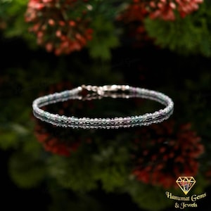 Puede incluir: Una delicada pulsera con pequeñas cuentas redondas multicolores en tonos verdes, morados y transparentes. La pulsera tiene un cierre plateado y se muestra sobre un fondo borroso rojo y verde.
