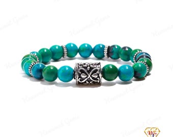 May include: A beaded bracelet featuring turquoise and green round beads. The bracelet has a decorative silver-colored cylindrical bead in the center. The beads are strung together with a stretch cord. The bracelet is a piece of jewelry.