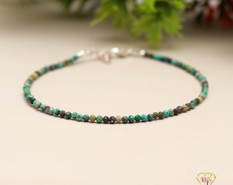 May include: A delicate beaded bracelet featuring small, round turquoise and brown beads. The beads are strung together with a silver-colored chain and clasp. The bracelet is displayed on a neutral surface, with greenery in the background.