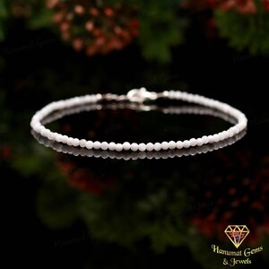 May include: A delicate white beaded bracelet with a silver clasp. The bracelet is composed of small, round beads and is displayed on a reflective surface. The background is blurred, with hints of green and red.