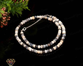 May include: A beaded necklace featuring small, round, multi-colored gemstones in shades of white, gray, and black. The necklace is strung on a silver chain and has a clasp closure. The background is black, with a sprig of greenery in the upper left corner.