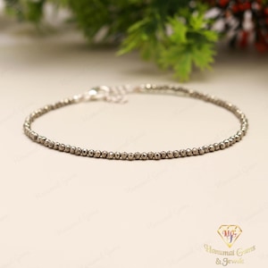 May include: A delicate silver bracelet featuring small, round, metallic beads strung together. The bracelet has a simple design with a clasp closure. The background includes greenery and a logo.