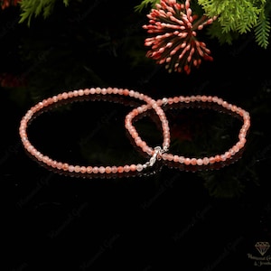 Può includere: Due delicati braccialetti realizzati con piccole perline rotonde color pesca. I braccialetti sono collegati da una chiusura argentata. Lo sfondo è nero, con vegetazione e un fiore rosso negli angoli superiori. I braccialetti sono per il polso.