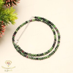 May include: A necklace with small, faceted beads in green, black, and red. It has a silver-coloured chain and clasp. The necklace is displayed on a cream-coloured surface. The beads are strung together.
