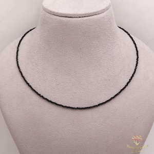 May include: A delicate black beaded necklace displayed on a mannequin. The necklace is composed of small, evenly spaced beads, creating a simple yet elegant design. The beads appear to be a deep black color, and the necklace is draped around the mannequin's neck.