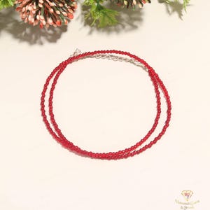 May include: A fine red beaded necklace with a silver clasp. The necklace is made of small, round beads and is shown on a light-coloured surface. The jewellery is a simple, elegant accessory.