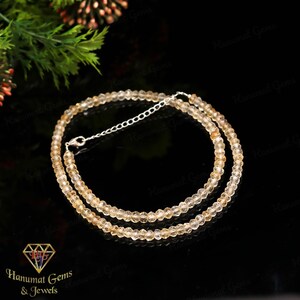 Peut inclure: Un collier délicat composé de petites pierres précieuses jaune clair et facettées. Le collier est doté d'une chaîne et d'un fermoir argentés. Le fond est noir, avec le texte "Hanumat Gems & Jewels" en bas à gauche.