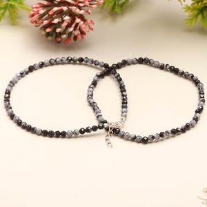 Può includere: Due cavigliere di perline con una miscela di pietre nere e grigie. Le cavigliere sono collegate da una chiusura color argento. Le perline, piccole e rotonde, creano un aspetto delicato. Presentate su una superficie chiara.