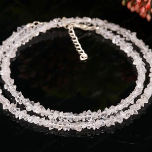 Pode incluir: Um colar delicado, em camadas, com vários fios de pedras preciosas claras e facetadas. O colar tem um fecho prateado e é exibido sobre um fundo escuro, realçando o brilho das pedras. As pedras preciosas parecem ser de corte bruto.