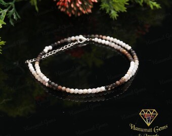 May include: A delicate necklace featuring two strands of small, faceted beads in shades of white, brown, and black. The necklace has a silver-colored clasp and chain. The jewelry is displayed on a reflective black surface.