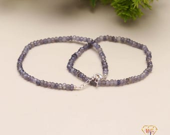 May include: Two necklaces made of small, faceted, blue-grey gemstones. The necklaces are connected by a silver clasp. The background is a light cream color with green foliage in the upper right corner.