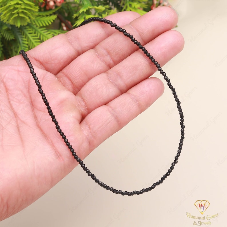 May include: A delicate black beaded necklace. The necklace is composed of small, round, black beads strung together. The necklace is displayed on a person's hand, with a soft, neutral background. The beads appear to be evenly spaced and have a subtle shine.