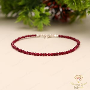 Peut inclure: Un bracelet délicat composé de petites pierres précieuses rondes, rouge foncé. Il est doté d'un fermoir argenté et est présenté sur une surface claire. Le fond comprend un feuillage vert et un logo. Le bracelet mesure environ 18 cm.