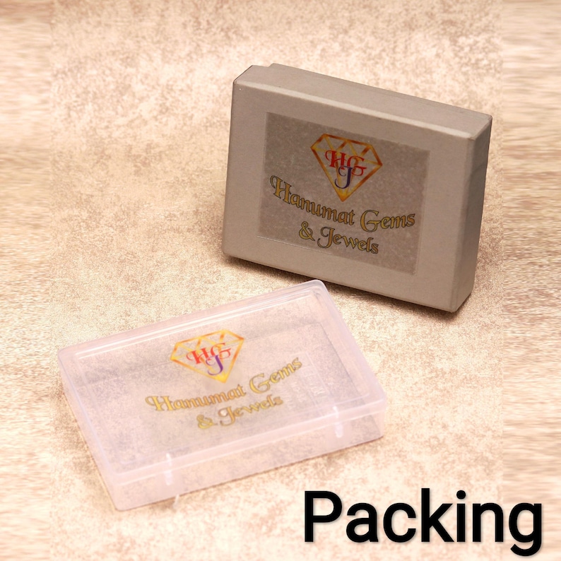 May include: Two jewelry boxes, one gray and one clear, are displayed. The gray box has a label with a logo and the text "Hanumat Gems & Jewels." The clear box is open, and the word "Packing" is written in black at the bottom.
