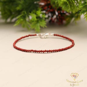 Peut inclure: Un bracelet délicat à perles rouges avec un fermoir argenté. Le bracelet est composé de petites perles rondes et est présenté sur une surface de couleur claire. Le fond présente un feuillage vert.