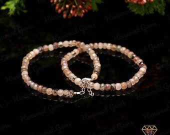 May include: Two beaded bracelets with a silver clasp. The bracelets are made of small, round, light brown and clear gemstones. The bracelets are displayed on a black surface with a blurred background of greenery and flowers.