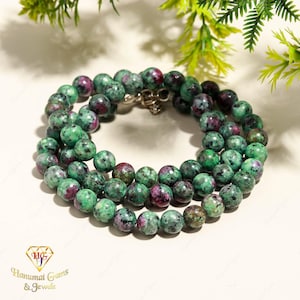 Può includere: Una collana di perline composta da pietre rotonde e levigate in tonalità di verde screziato e magenta intenso. Le perline sono infilate in una disposizione circolare e fissate con una chiusura in metallo argentato. Il logo Hanumat Gems & Jewels è visibile.