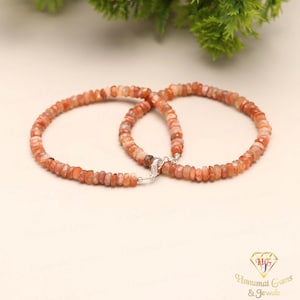 Puede incluir: Dos pulseras de piedras preciosas de color naranja, unidas por un cierre plateado. Las cuentas son pequeñas y de forma irregular. El fondo es de un color neutro, con un poco de follaje verde en la esquina superior derecha.