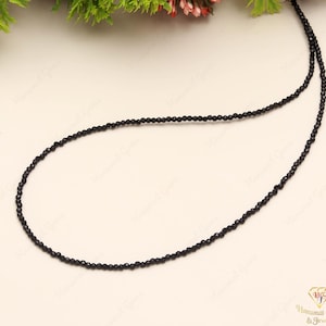 May include: A delicate black beaded necklace. The necklace is composed of small, round, black beads strung together to form a single strand. The beads are evenly spaced and create a simple, elegant design. The necklace is displayed on a light-colored surface.