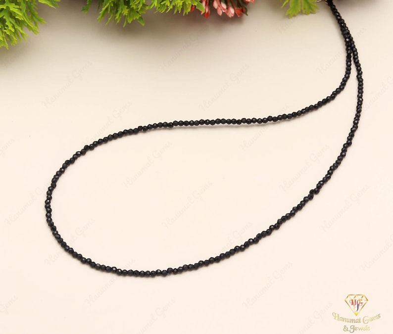 May include: A delicate black beaded necklace. The necklace is composed of small, round, black beads strung together to form a single strand. The beads are evenly spaced and create a simple, elegant design. The necklace is displayed on a light-colored surface.