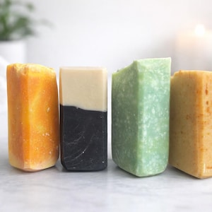 May include: Four rectangular bars of soap standing upright. The first is a vibrant orange, the second is split horizontally between cream and black, the third is a textured pale green, and the fourth is a speckled light tan with a rough surface.