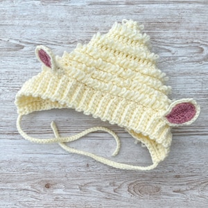 May include: A cream-colored, crocheted baby hat shaped like a lamb. The hat features two small, pink-lined ears and ties under the chin. The hat is made of soft yarn and has a textured, fluffy appearance, perfect for a baby.