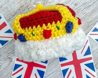 May include: A handmade crochet crown in yellow, red, and blue, with a white fluffy trim. The crown is decorated with red and blue accents. Union Jack flags are placed around the crown. The crown is likely a decorative item or a costume accessory.