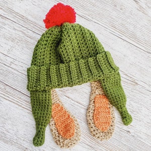 May include: Green crocheted hat with a red pom-pom and two long, crocheted ear flaps. The ear flaps are a light brown color.
