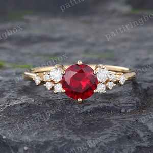 May include: A gold ring featuring a large red ruby gemstone surrounded by smaller white diamonds. The ring is displayed on a textured grey surface.