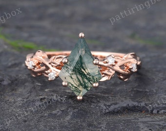 Puede incluir: Un anillo de compromiso de oro rosa con una piedra central de ágata musgosa y acentos de diamantes. La banda es un delicado diseño de vid trenzada.