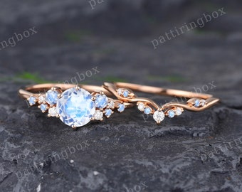 May include: A rose gold engagement ring set with a large round moonstone center stone and smaller blue and white accent stones. The band is adorned with delicate vine-like details.