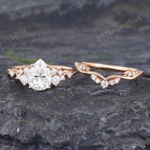 以下が含まれることがあります： ローズゴールドの婚約指輪セット。メインリングは、洋梨型のセンターストーンと、小さな石が散りばめられた曲線的なバンドが特徴です。セットは暗い表面に表示されています。
