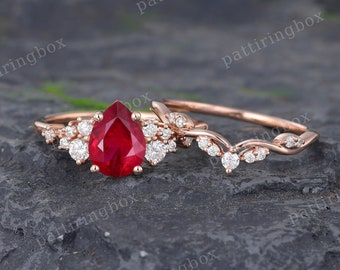 May include: A pear-shaped ruby engagement ring set in rose gold with a delicate diamond band. The ruby is surrounded by smaller diamonds, creating a sparkling halo effect.