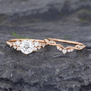Puede incluir: Un juego de anillos de compromiso de oro rosa con una piedra preciosa grande, redonda y transparente y piedras de acento más pequeñas. La banda a juego tiene un diseño curvo con pequeñas piedras preciosas. Los anillos se muestran sobre una superficie oscura y texturizada.