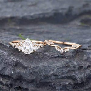 以下が含まれることがあります： ローズゴールドの婚約指輪セット。ペアシェイプのセンターストーンを小さなラウンドダイヤモンドが囲み、それに合わせたカーブを描く結婚指輪には、葉のような形の繊細なマーキスカットとラウンドカットのダイヤモンドがあしらわれています。