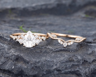 May include: A rose gold engagement ring set featuring a pear-shaped center stone surrounded by smaller round accent stones, paired with a matching curved wedding band adorned with a series of small, delicate round stones along the band.