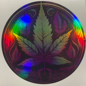 May include: A round, iridescent sticker with a black background and a detailed, stylized image of a marijuana leaf in the center. The leaf is surrounded by smaller leaves and swirls, and the sticker has a gold border.