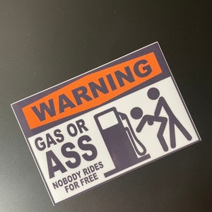 May include: A white sticker with black and orange text that reads "WARNING GAS OR ASS NOBODY RIDES FOR FREE". The sticker also features a black silhouette of a gas pump and two stick figures.