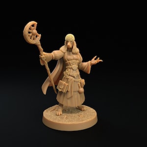 Puede incluir: Una figura en miniatura de color marrón claro de un personaje de fantasía, posiblemente un mago o hechicera, sosteniendo un bastón con un diseño de media luna. La figura lleva una túnica larga y tiene el pelo largo. La figura está sobre una base circular.