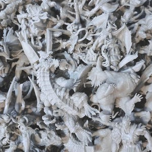 Puede incluir: Primer plano de una colección de pequeñas figuras de fantasía grises sin pintar. Las figuras incluyen dragones, monstruos y otras criaturas míticas. Las figuras están densamente empaquetadas, creando una superficie texturizada. La impresión general es de un mundo de fantasía en miniatura.