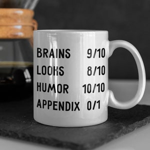May include: White ceramic mug with black text that reads "Brains 9/10, Looks 8/10, Humor 10/10, Appendix 0/1".