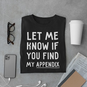 May include: Black t-shirt with white text that reads "Let me know if you find my appendix". The shirt is folded and laid flat on a gray surface with a coffee cup, a smartphone, a pair of glasses, and a notebook and newspaper.