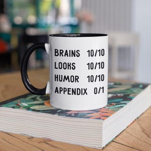 May include: White coffee mug with a black handle and black rim. The mug has black text that reads "Brains 10/10, Looks 10/10, Humor 10/10, Appendix 0/1".