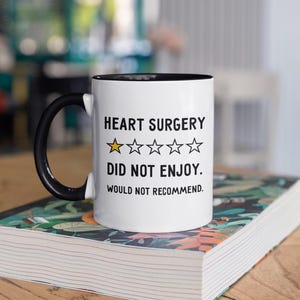 Pu&ograve; includere: Una tazza da caff&egrave; bianca con bordo e manico neri. La tazza ha un testo nero che recita "HEART SURGERY" con una valutazione di una stella e il testo "DID NOT ENJOY. WOULD NOT RECOMMEND."