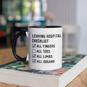 May include: White ceramic mug with a black handle and black rim. The mug has the text "Leaving Hospital Checklist" with checkboxes and the text "All Fingers", "All Toes", "All Limbs", and "All Organs".