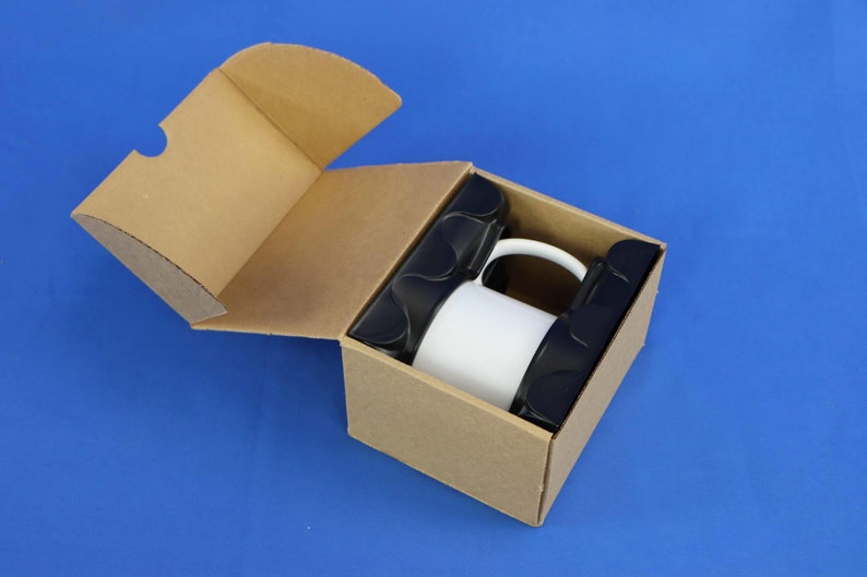 Pu&ograve; includere: Una tazza in ceramica bianca &egrave; all'interno di una scatola di cartone marrone con un inserto in schiuma nera. La scatola &egrave; aperta e la tazza &egrave; visibile.