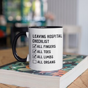 May include: White ceramic mug with a black handle and black rim. The mug has a checklist on it that says "Leaving Hospital Checklist" with checkboxes next to the following items: "All Fingers", "All Toes", "All Limbs", and "All Organs". The mug is sitting on a stack of books.