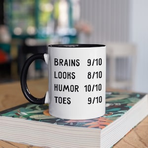 May include: White ceramic mug with a black handle and black rim. The mug has black text that reads "BRAINS 9/10 LOOKS 8/10 HUMOR 10/10 TOES 9/10". The mug is sitting on a book with a colorful floral design.