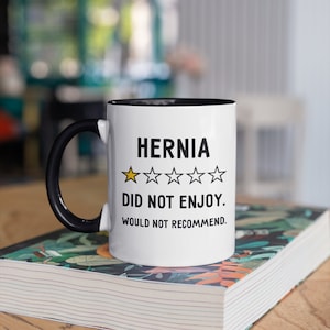 May include: White ceramic mug with a black handle and black rim. The mug has a black graphic that says "Hernia" with a one-star rating and the text "Did not enjoy. Would not recommend."